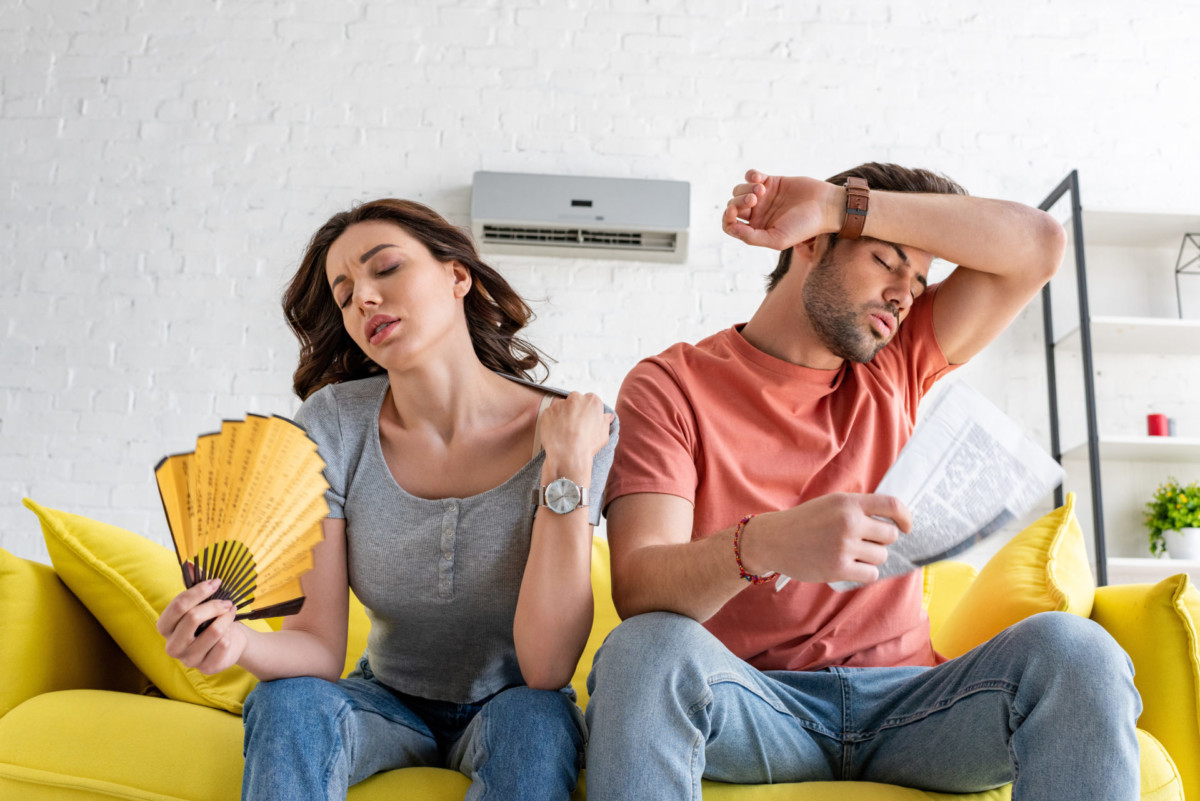 A man and a woman sitting on a yellow couch look uncomfortable due to heat, using paper fans and a magazine to cool off, with an air conditioner in the background.