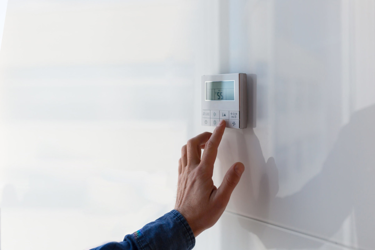 A person adjusting a digital thermostat on a white wall, with sunlight streaming through a window nearby.