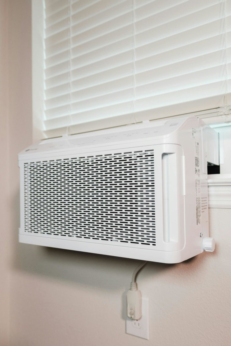 A white window air conditioner installed under a closed window with blinds, with its power cord plugged into a nearby wall outlet.