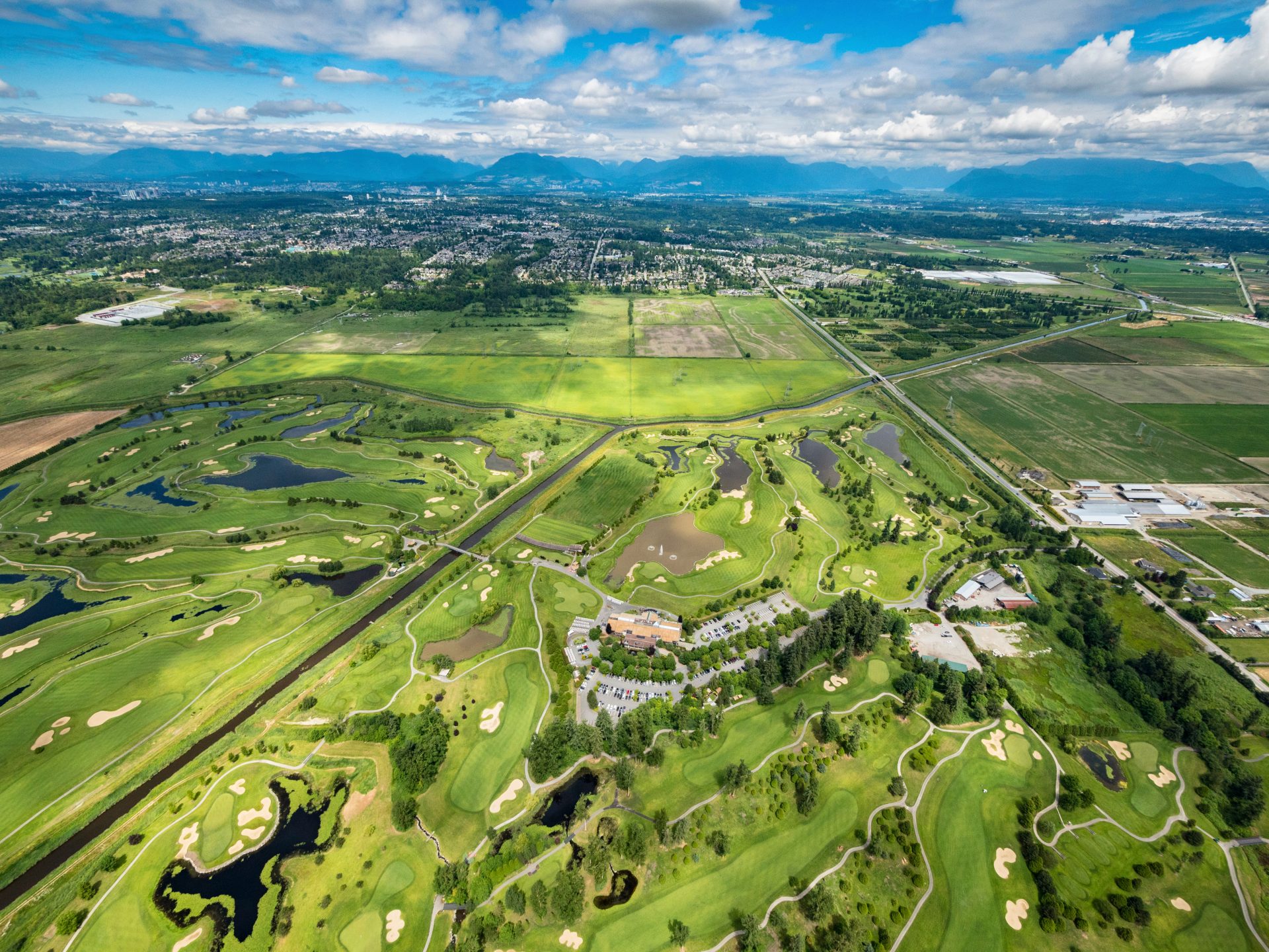 Aerial view of an expansive golf course with greens, water features, pathways, a central clubhouse, and surrounding open fields under a partly cloudy sky.