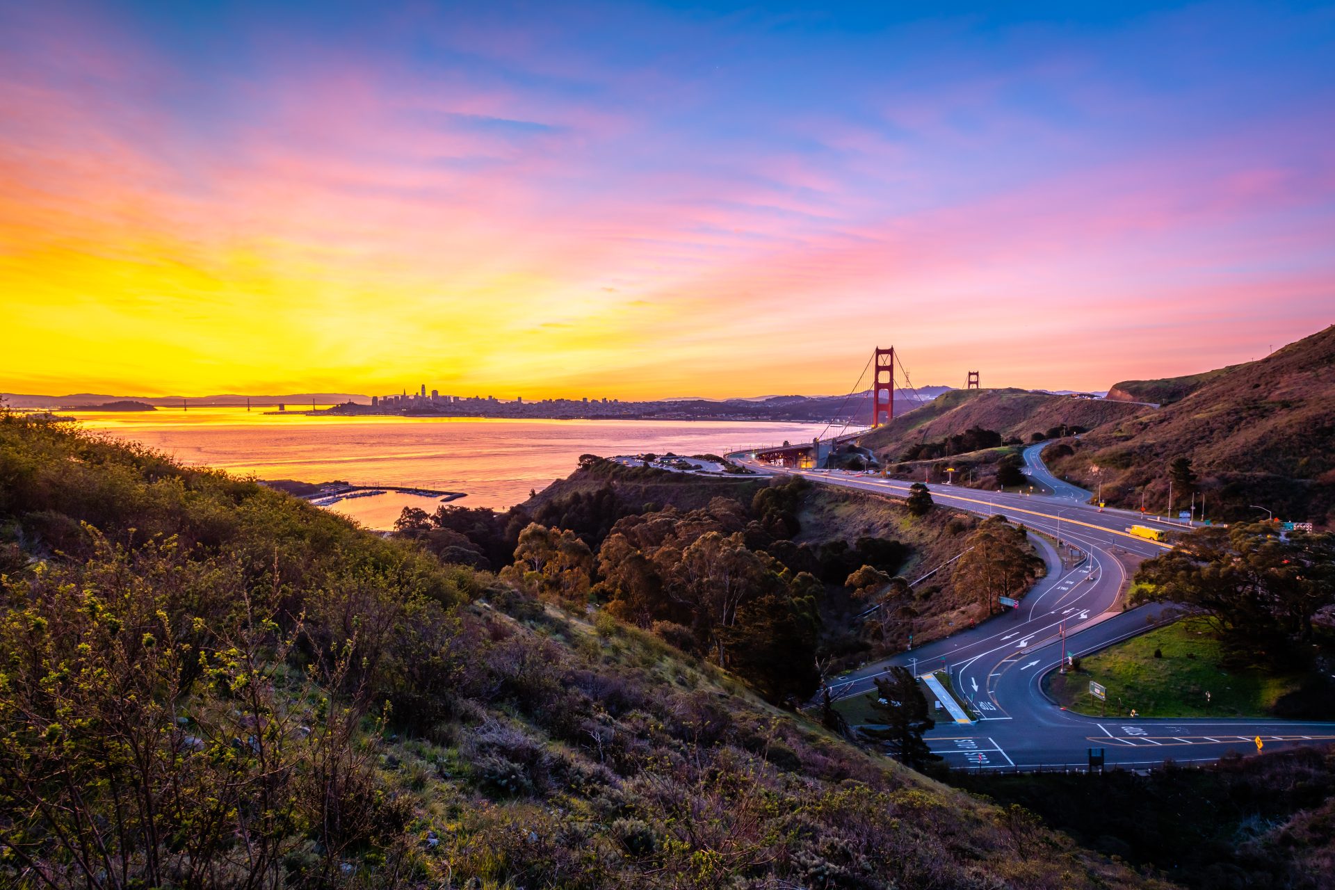 The Golden Gate Bridge at sunset with the San Francisco skyline in the distance, winding roads and Marin County hills in the foreground.