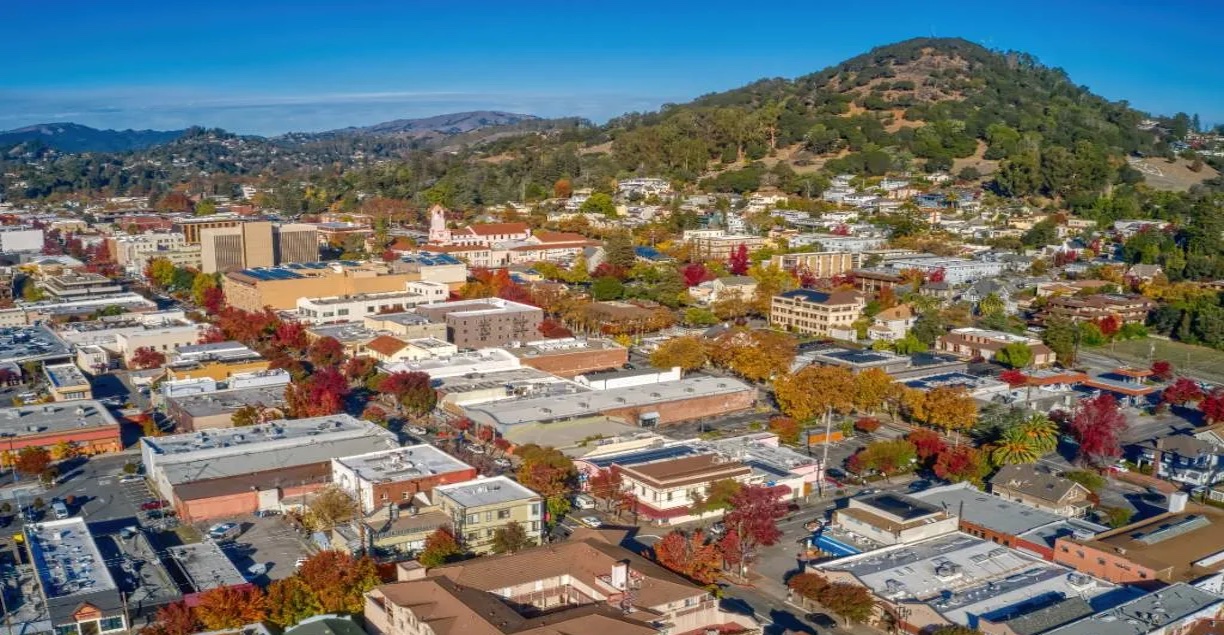 Aerial view of a small city with a mix of buildings, colorful trees, and a wooded hill in the background under a clear blue sky.