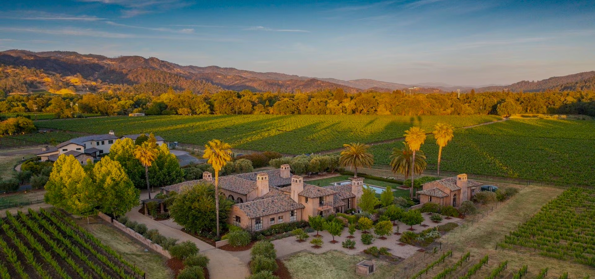 Aerial view of a large house with a tile roof surrounded by vineyards, trees, and mountains under a clear sky at sunset.