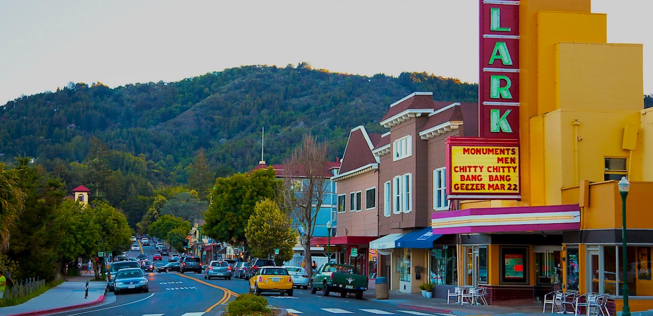 A small downtown street with cars parked along the side and a vintage theater marquee reading movie titles, with green hills in the background.