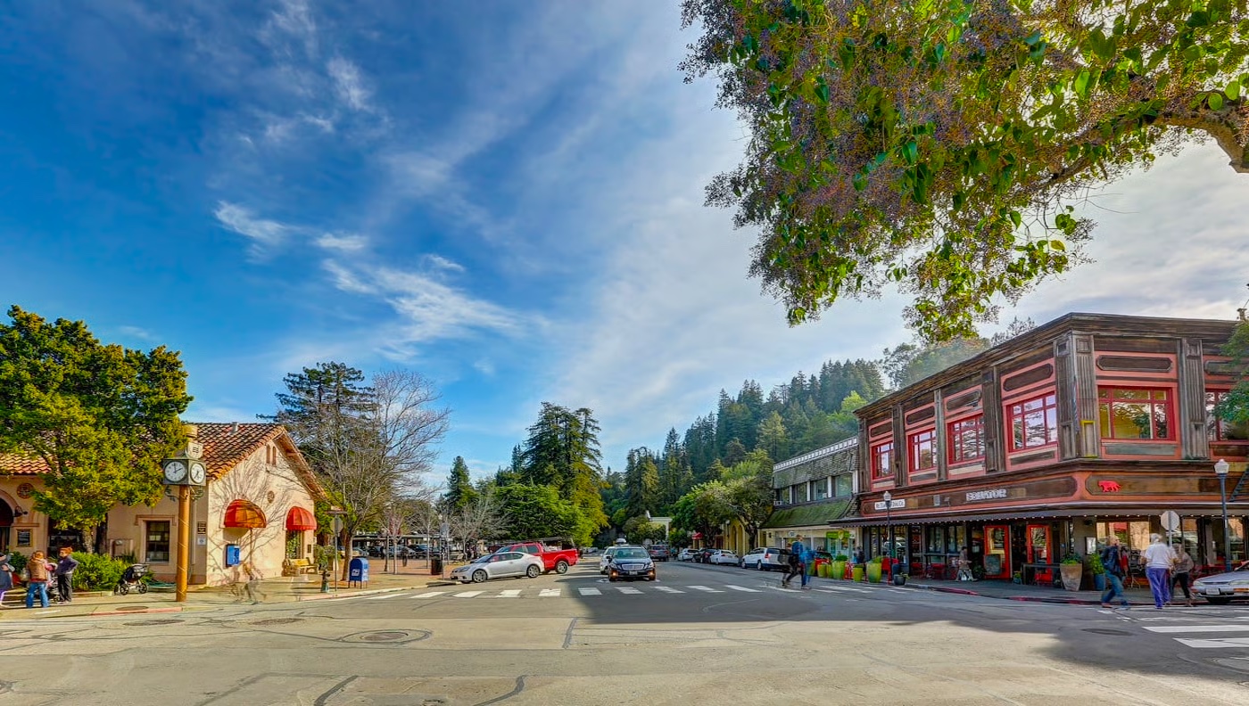 A street intersection in a small town with shops, parked cars, people walking, and trees under a partly cloudy sky.