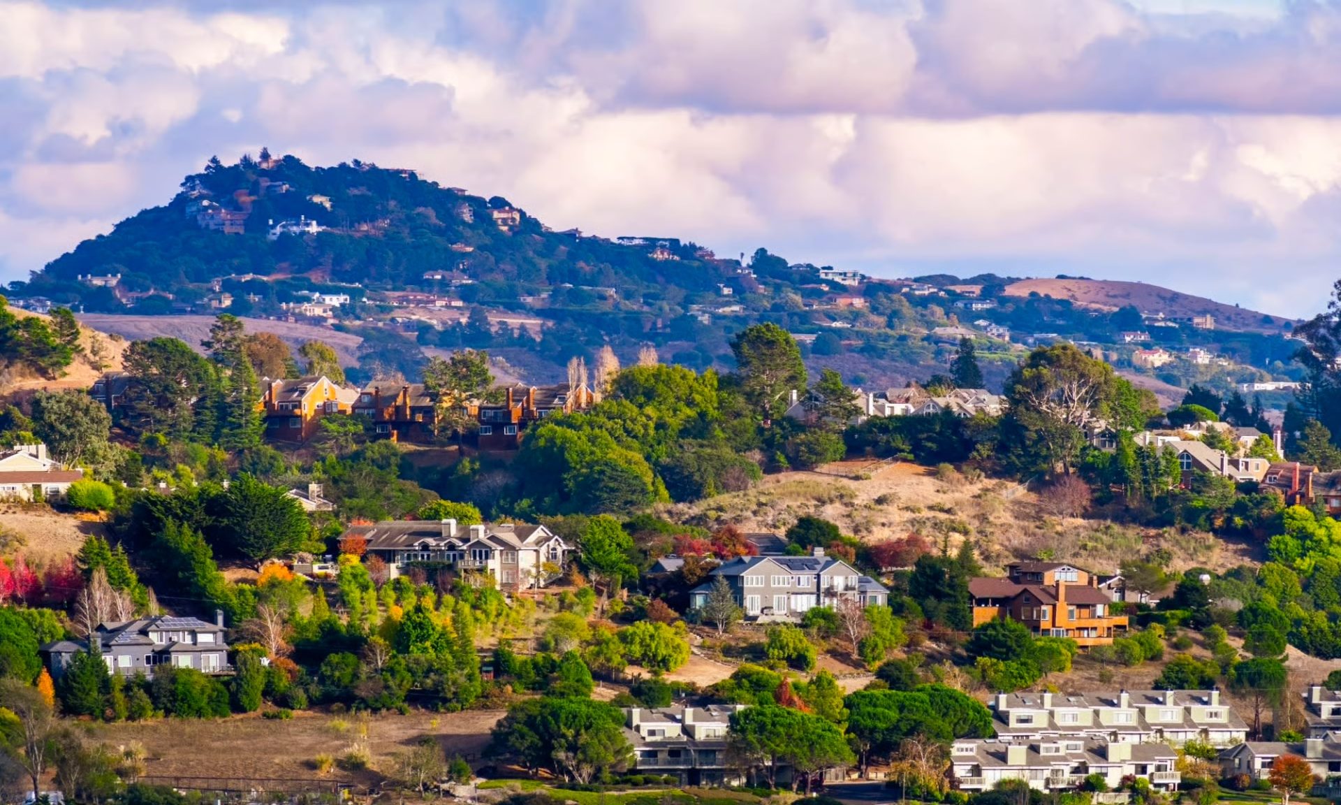 Houses are scattered across hilly terrain with a large, tree-covered mountain in the background under a partly cloudy sky.