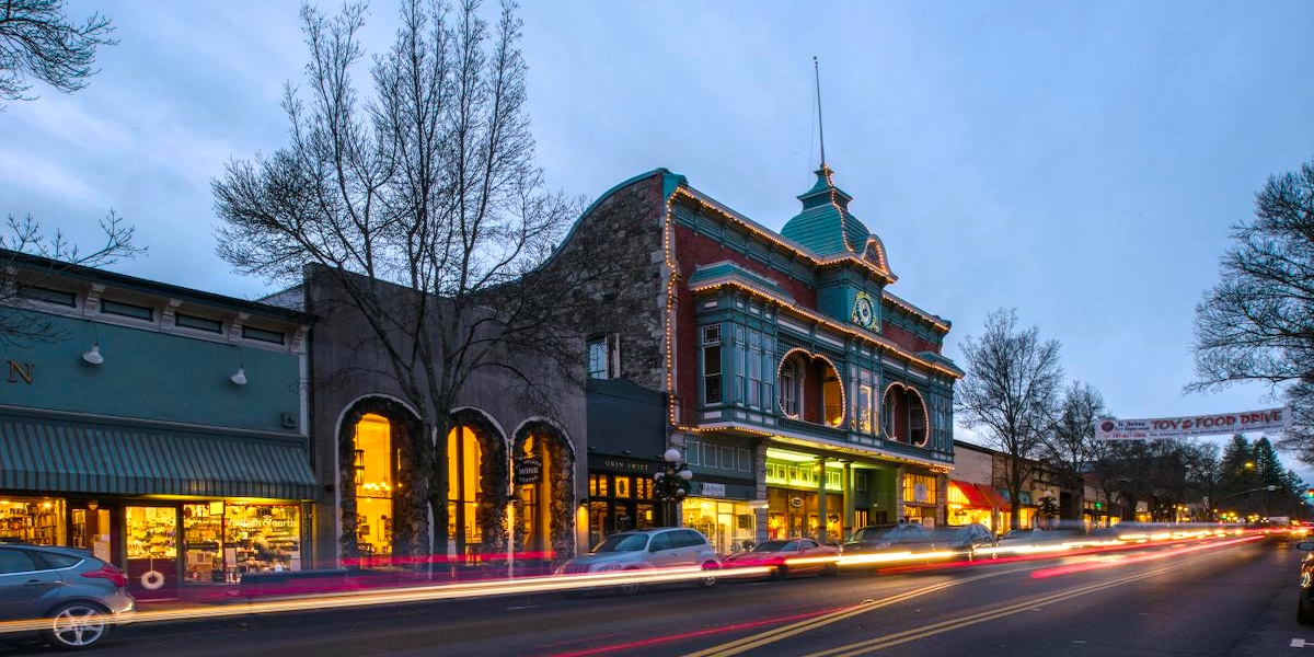 A street view at dusk shows historic buildings with storefronts, light trails from passing cars, and leafless trees lining the sidewalk.