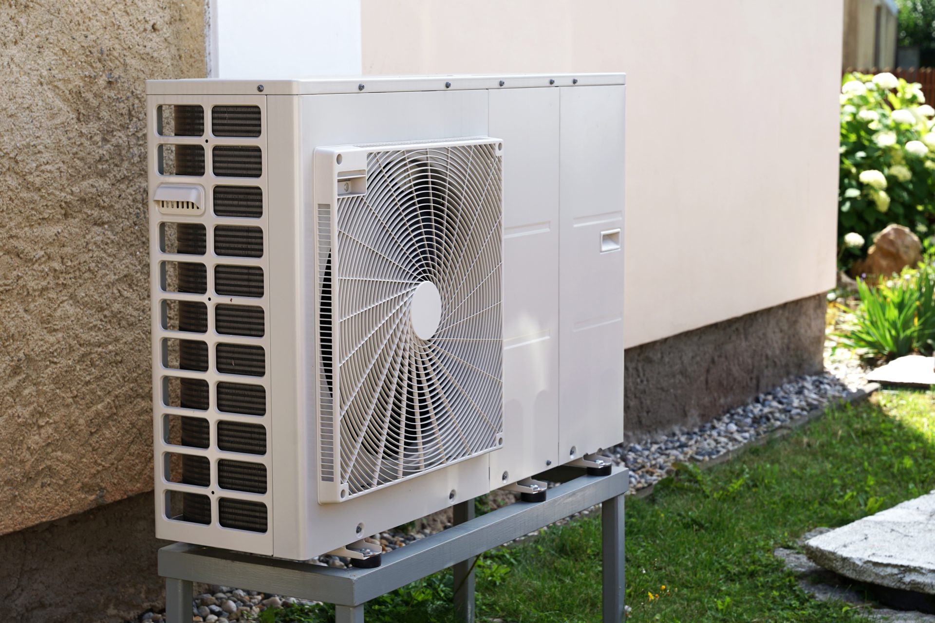 A white outdoor heat pump unit is mounted on a metal stand next to a home, surrounded by grass and landscaping stones in Northern California.