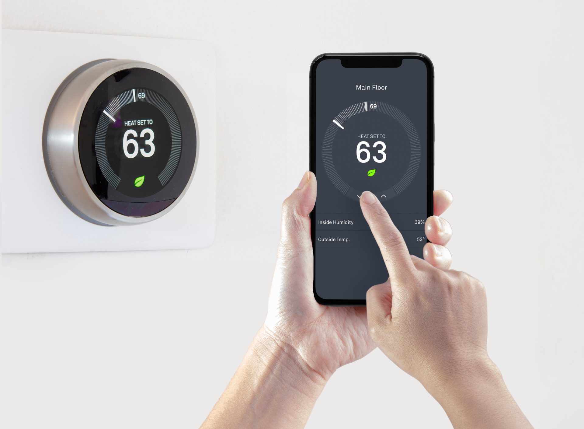A person adjusts a smart thermostat set to 63°F using a smartphone app, aiming to lower energy bills. The thermostat is mounted on a white wall.