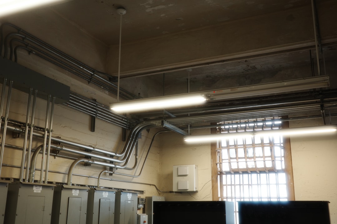 Fluorescent lights and exposed electrical conduits run along the ceiling of an industrial room with a barred window and multiple electrical panels, highlighting the importance of regular HVAC maintenance in such environments.