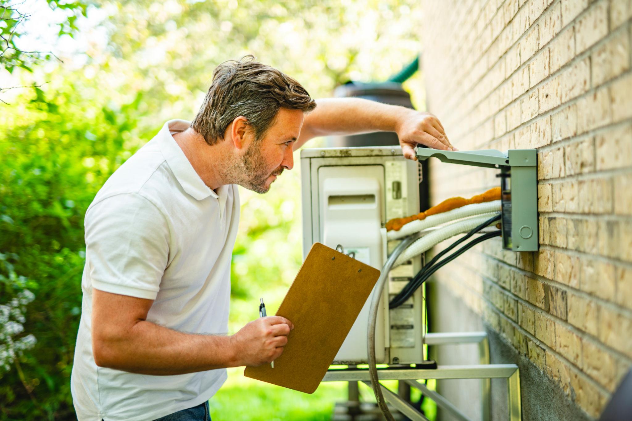 A man holding a clipboard inspects an outdoor air conditioning unit mounted on a brick wall, ensuring the Santa Rosa Air Conditioner is ready for the summer heat. A man holding a clipboard inspects an outdoor air conditioning unit mounted on a brick wall, ensuring the Santa Rosa Air Conditioner is ready for the summer heat.