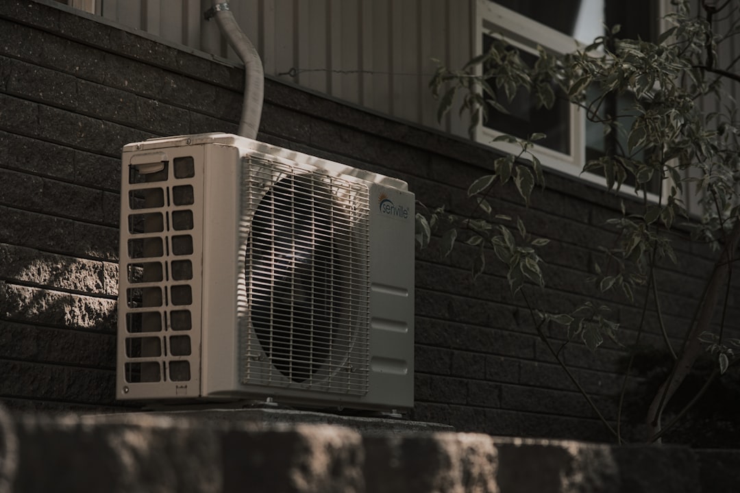 An outdoor air conditioning unit is installed against a dark brick wall beneath a window, with a plant nearby—typical of professional Furnace Installation or Heat Pump Installation services often seen in Marin and Sonoma County.